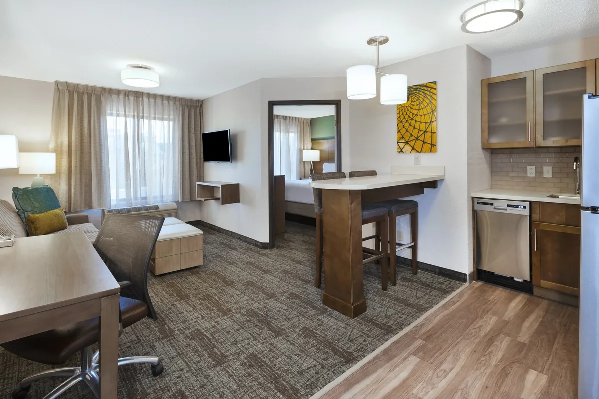 The hero image features a spacious, contemporary living area with plush seating, a sleek kitchenette, dedicated workspace, and tasteful artwork, all bathed in warm, inviting light. The layout invites guests to unwind, work remotely, or gather for intimate meals in comfort and style.