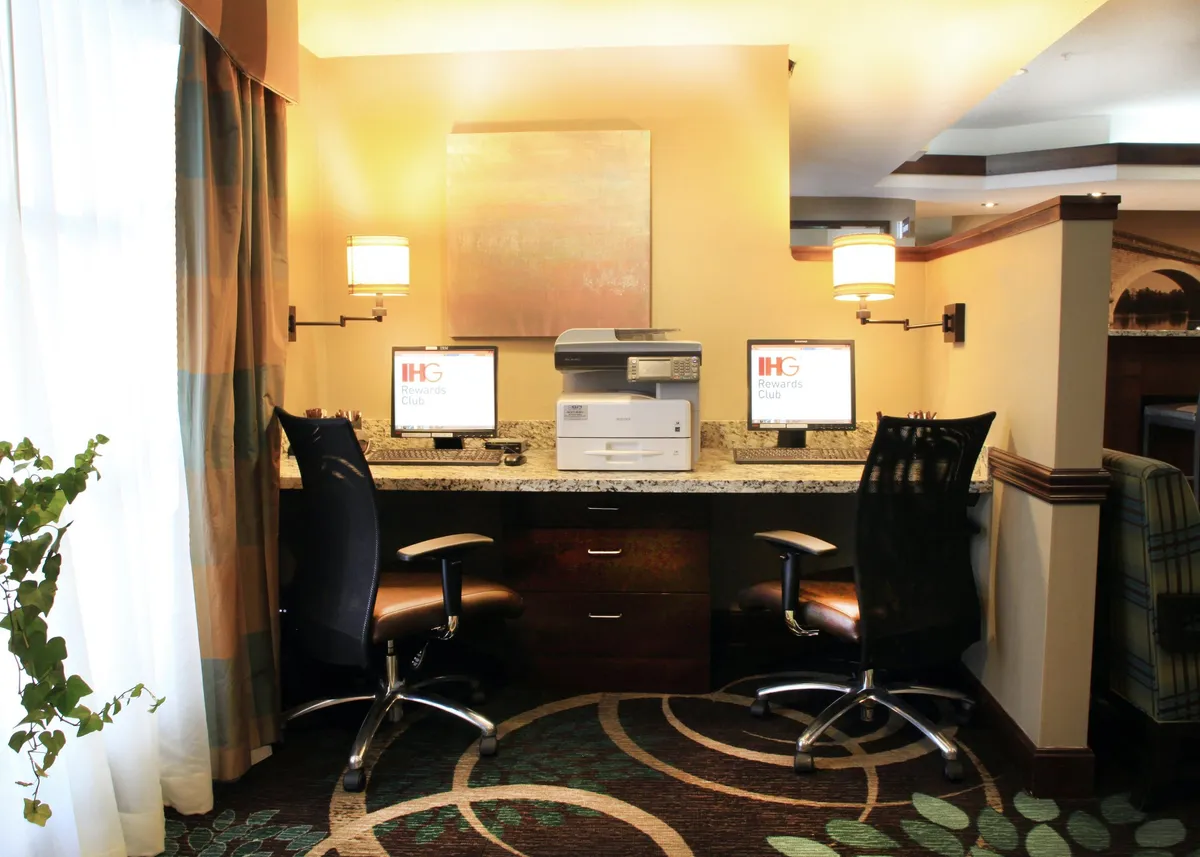 A sleek business center featuring two computer desks equipped with monitors, a printer, modern lighting, and an art piece on the wall. The workspace is organized and well-lit, creating a professional environment ideal for focused work and productivity.