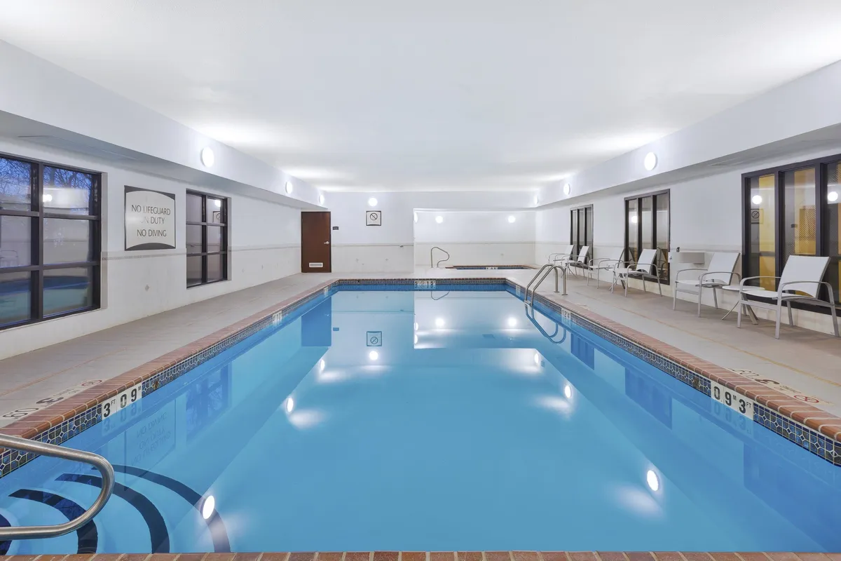 A serene indoor swimming pool area with large windows allowing natural light to flood the space. Lounge chairs line the pool, inviting guests to relax and socialize in a calm, airy environment.