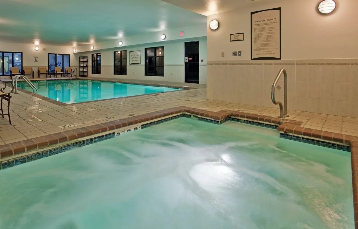 A bright, tranquil indoor pool space featuring both a generously sized swimming pool and an adjacent bubbling hot tub. There are several comfortable seating areas, and the ambiance is serene and inviting, perfect for relaxation and social gatherings.