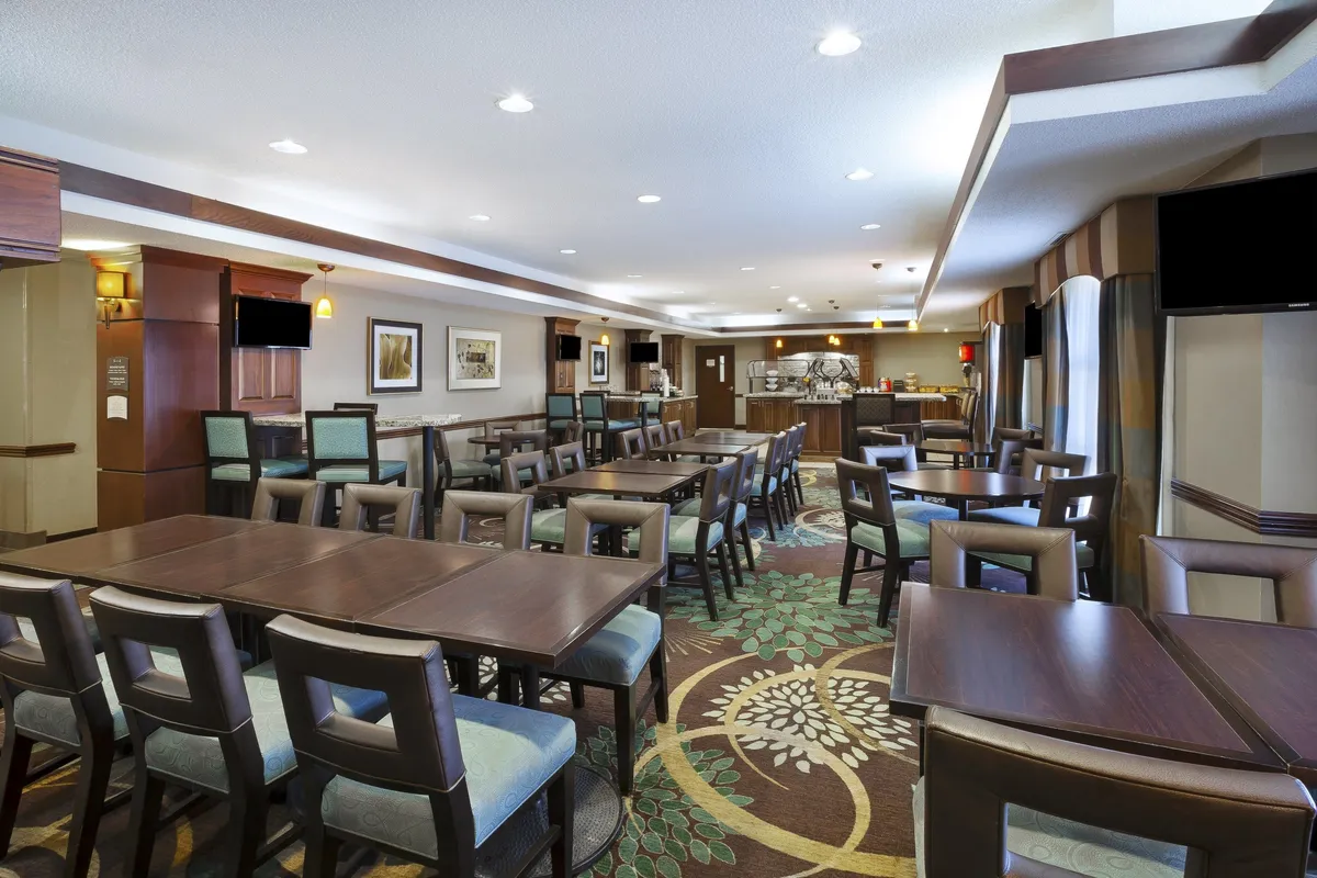 A spacious dining area with wooden tables, upholstered chairs, decorative carpet, wall art, and a visible buffet area. The setting is inviting and suitable for both meals and social gatherings.