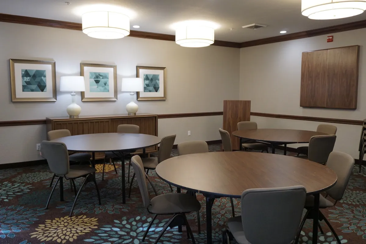 A comfortable meeting or workshop room is shown, with round tables, decorative lamps, framed artwork, and patterned carpet. The setting accommodates both professional meetings and casual gatherings in a comfortable atmosphere.