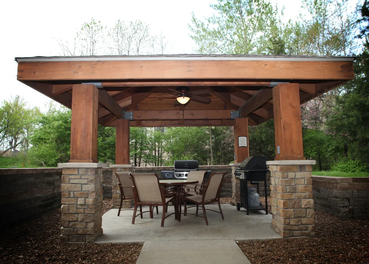 A welcoming outdoor dining area featuring a large table, a grill, ceiling fan, and sturdy stone pillars. The space is shaded and well-equipped for gatherings, with plenty of room for families to sit and enjoy meals together. The setting hints at the possibility of sunset dinners, morning coffee outdoors, or casual family picnics.